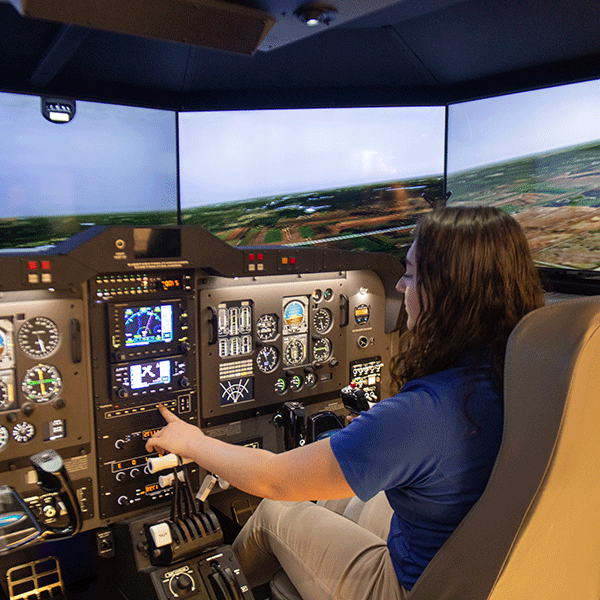 A student with dark hair, wearing a blue shirt and beige pants, works the controls of a flight simulator. An aerial scene is visible on the three simulation screens. The simulator control panel features switches, buttons, gauges, and digital screens.